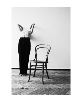 Ilya, photographed by Lilit L Hovhanni — standing beside a bentwood chair, face hidden behind raised arms.